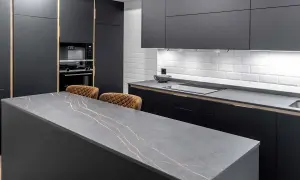 Isla de cocina moderna con encimera de piedra gris veteada en oro. Dos taburetes de cuero marrón esperan junto a la medida encimera.
