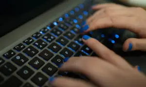 Dłonie z niebieskim lakierem na paznokciach piszą na podświetlanej klawiaturze MacBooka. Twarda spacja jest widoczna.