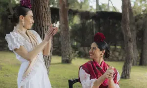 Dos mujeres vestidas para ir al Rocío, con trajes blancos y mantones, sonríen y aplauden en un jardín.