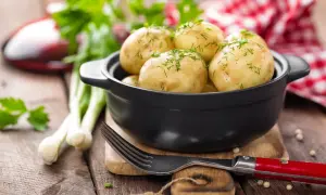 Młode ziemniaki w czarnej misce, posypane koperkiem, obok widelca i dymki.