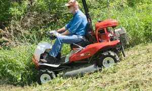 Mężczyzna w czapce i okularach prowadzi kosiarkę Brush Rover, kosząc wysokie trawy. Dobre opinie o dedra sprzęcie potwierdzają jego skuteczność.