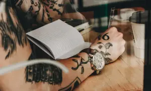 Dłoń z tatuażem na mostku pisze w notatniku. Na nadgarstku zegarek.