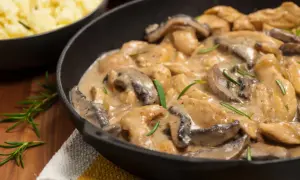 Pyszny kurczak w sosie pieczarkowym, podany w żeliwnej patelni, z gałązkami rozmarynu. Obok widoczna miska z purée ziemniaczanym.