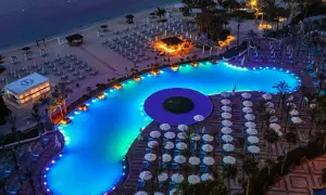 Nocny widok na luksusowy basen i plażę w Dubaju. Błękitna woda basenu, oświetlona kolorowymi światłami, otoczona leżakami i palmami.