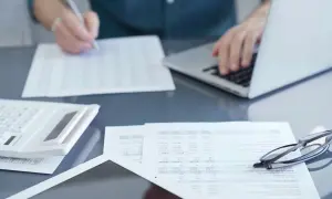 Osoba pracuje przy laptopie, przeglądając dokumenty i pisząc. Na biurku leży kalkulator i okulary. Przygotowania do podpisania umowy zlecenie 2026.