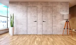 Dwa minimalistyczne, **drzwi ukryte w ścianie** z betonu architektonicznego, z czarnymi uchwytami. Obok stoi lampa na trójnogu.