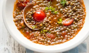 Una ciotola bianca piena di saporita zuppa di lenticchie con pomodorini e prezzemolo fresco. Un mestolo serve la zuppa.
