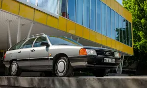 En grå Audi 100 C3 Avant står parkerad framför en modern gul byggnad med stora fönster.