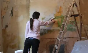 Kobieta z pędzlem w ręku usuwa tynk strukturalny ze ściany. Obok stoi drabina.