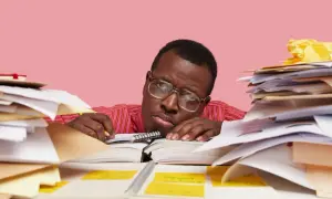 Un homme fatigué, entouré de piles de documents, semble dépassé par sa tâche. Il est peut-être en train de préparer un préavis foncia.