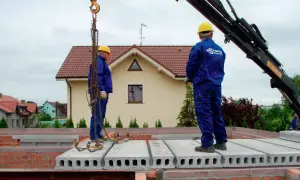 Dwóch budowlańców w żółtych kaskach montuje prefabrykowane płyty stropowe.