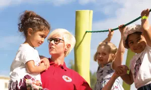Frau vom Roten Kreuz fördert **Inklusion im Kindergarten**, indem sie ein Kind hält, während andere Kinder an einem Seil spielen.