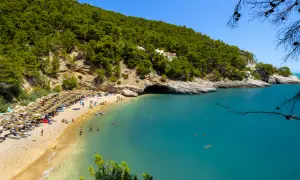 Spiaggia affollata con ombrelloni, gente che nuota nel mare turchese e una collina verdeggiante. Un pugno chiuso di natura incontaminata.