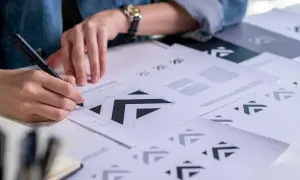 Osoba szkicuje logo wektorowe na papierze, otoczona wieloma wydrukami tego samego projektu.