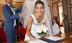 La mariée Anne-Claire Legendre signe son acte de mariage, bouquet à la main. Un enfant aux boucles blondes observe la scène.