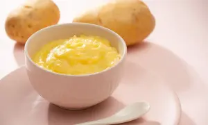 Ein Schälchen mit cremigem Kartoffelbrei, bereit für ein Baby. Zwei Kartoffeln liegen im Hintergrund.