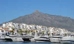 Luksusowe jachty cumują w malowniczym porcie Puerto Banús, z białymi budynkami i górą w tle.