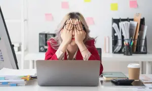 Kobieta w biurze z rękami na twarzy, wyraźnie zmęczona. Widoczne objawy stresu długotrwałego: stosy papierów, laptop, kubek kawy.