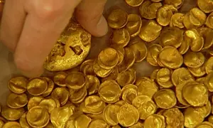 Ein Finger berührt einen Teil des Goldschatzes von Manching, der aus vielen kleinen, unregelmäßigen Goldmünzen besteht.