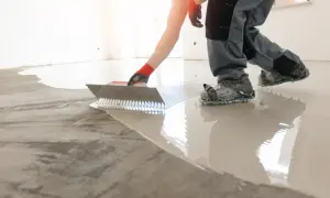 Obrero con guantes rojos y calzado especial nivela un suelo con mucho desnivel usando una llana dentada.