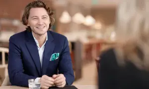 Rasmus Nerman, klädd i en mörkblå kavaj, ler och tittar åt sidan under ett samtal vid ett bord.