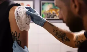 Artysta w rękawiczkach dopasowuje wzór tatuażu na ramieniu klienta. Widać już kilka gotowych tatuaży, co sugeruje, że to miejsce na tatuaż jest popularne.