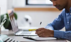 Mężczyzna notuje, zastanawiając się, jak sprawdzić, co spowalnia komputer. Na biurku laptop i notatnik.