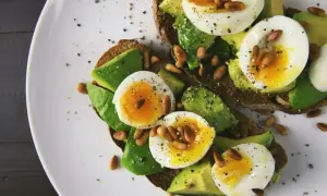 Avokado recept: två rostade brödskivor med avokado, kokt ägg och pinjenötter, strösslade med peppar.