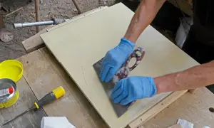 Händer med blå handskar skrapar bort slipdamm från en ljusgul yta med en bred spackel. Viktigt att få bort slipdamm innan målning för ett perfekt resultat.