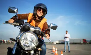 Uśmiechnięta kursantka w kamizelce i kasku na motocyklu, gotowa do zdobycia prawa jazdy na motocykl. Instruktor obserwuje w tle.