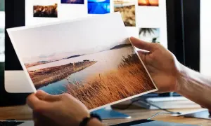 Dłonie trzymają wydruk krajobrazu. Różne rodzaje papieru do druku zdjęć są widoczne na ekranie komputera w tle.