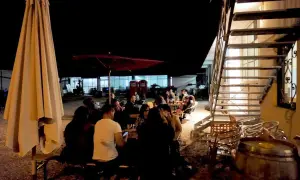 Serata al birrificio Mastro Matto: gente seduta ai tavoli sotto un grande ombrellone, con una scala metallica sullo sfondo.