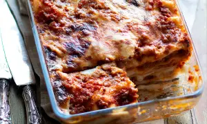 Lasagna al ragù appena sfornata, dorata e filante, in una teglia di vetro, con posate d'argento a lato.