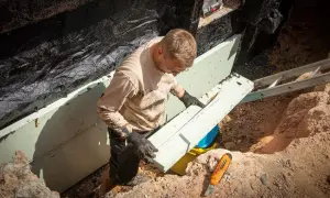 Mann verlegt Noppenbahn zur Kellerwand Abdichtung. Der Bauarbeiter trägt Handschuhe und eine Schutzbrille.