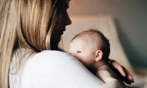 En mamma håller sitt barn i famnen, med barnet vänt mot hennes axel. Det ser ut som ett ögonblick av lugn och närhet, kanske under amning med ett klassiskt hamburgergrepp.