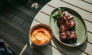 En skål med krämig röra, troligen med creme fraiche, fetaost och soltorkade tomater, bredvid en tallrik med grillad fisk.