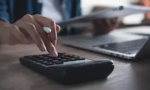 Ręka na kalkulatorze, obok laptop i dokumenty. Obliczanie, ile trzeba przepracować, by dostać 13 pensję.