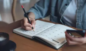 Dłoń pisze w planerze, planując swój staz pracy. Obok filiżanka kawy i telefon.