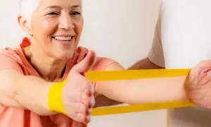 Uśmiechnięta starsza kobieta ćwiczy z fizjoterapeutą, używając żółtej gumy rehabilitacyjnej.