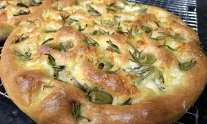 Une focaccia pizza dorée, garnie d'olives vertes et de romarin frais, sort du four.