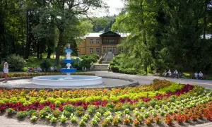 Uzdrowisko z fontanną i kolorowymi rabatami kwiatowymi przed budynkiem sanatorium.