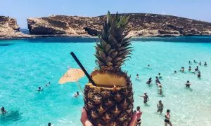 Koktajl z ananasa na plaży na Malcie. Woda jest turkusowa, a na horyzoncie widać skaliste klify. Ceny na Malcie są przystępne.