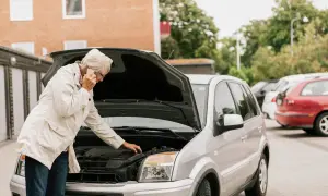 Äldre kvinna med telefon vid öppen motorhuv på en silverfärgad bil. Hon verkar orolig och undrar om man kan besikta bilen tidigare.
