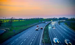 Autostrada z ruchem samochodowym o zachodzie słońca, z zielonymi polami w tle.
