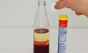 Eine Hand lässt eine Tablette in eine Flasche mit bunten Flüssigkeiten fallen, um eine Lavalampe selber zu machen.