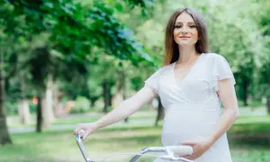 Uśmiechnięta ciężarna kobieta w białej sukience trzyma kierownicę roweru w parku. Czy w ciąży można jeździć na rowerze? Tak, jeśli czujesz się dobrze!