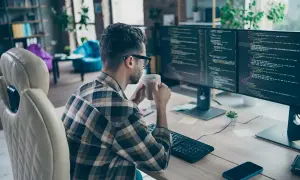 Programista w okularach pije kawę, pracując zdalnie przy dwóch monitorach z kodem.