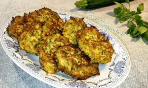 Frittelle di zucchine dorate e croccanti, servite su un piatto decorato. Accanto, una zucchina fresca e un rametto di erbe aromatiche.