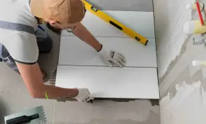 Un albañil coloca baldosas blancas sobre yeso, usando un nivel y un láser para asegurar una instalación perfecta.