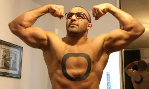 Muskularny mężczyzna o arabskich rysach, w okularach, prezentuje swoje bicepsy. Na klatce piersiowej ma tatuaż w kształcie pierścienia.